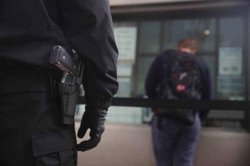 A security officer in a black uniform with a handgun holstered at his side stands in the foreground, while a person in the background is leaning over a counter.
