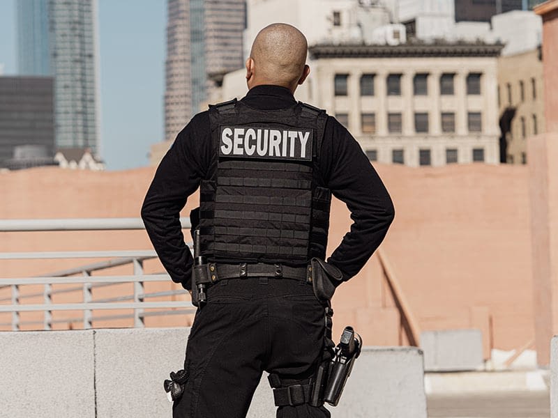 A security officer wearing a uniform with 'SECURITY' emblazoned on the back, standing on a rooftop overlooking a cityscape.